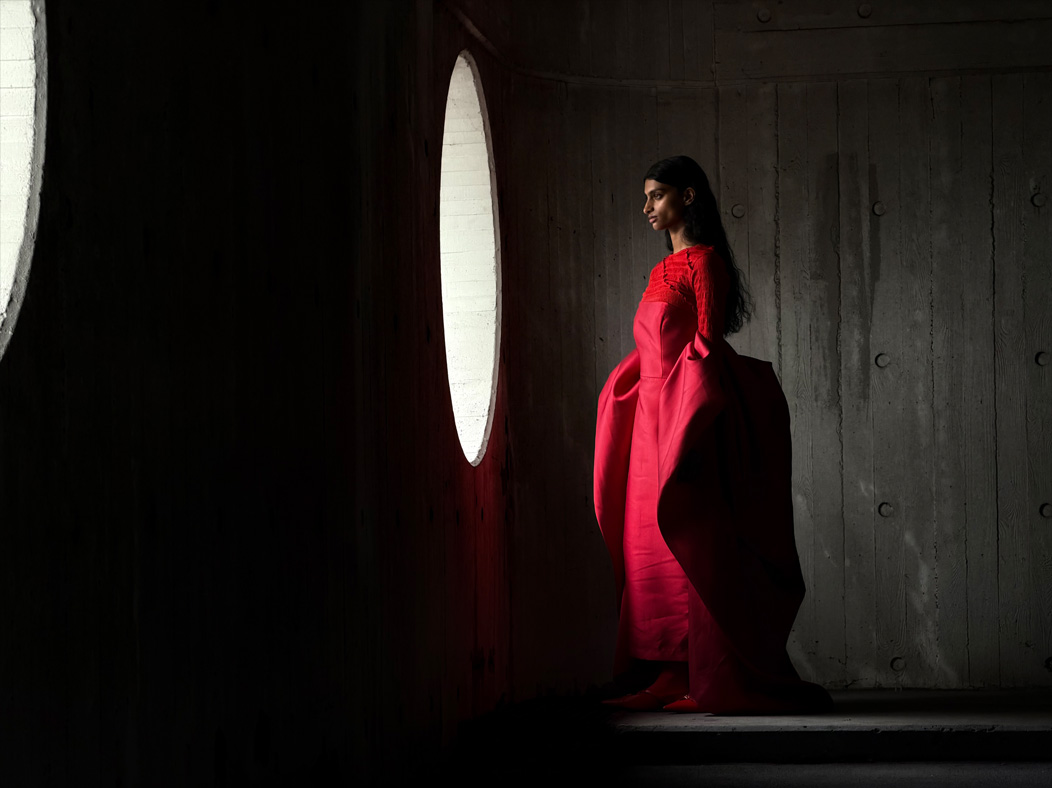 Una donna con un lungo vestito rosso fotografata in piedi in una stanza vuota. Nell’app Foto la lunghezza focale viene regolata a 100 mm, per mettere in evidenza le capacità del sistema di fotocamere Pro